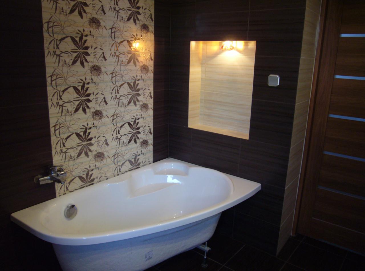 Biała wanna narożna na tle ciemnobrązowych płytek z kwiatowym wzorem i podświetlonej wnęki, drewniane drzwi z przeszkleniami po prawej stronie.