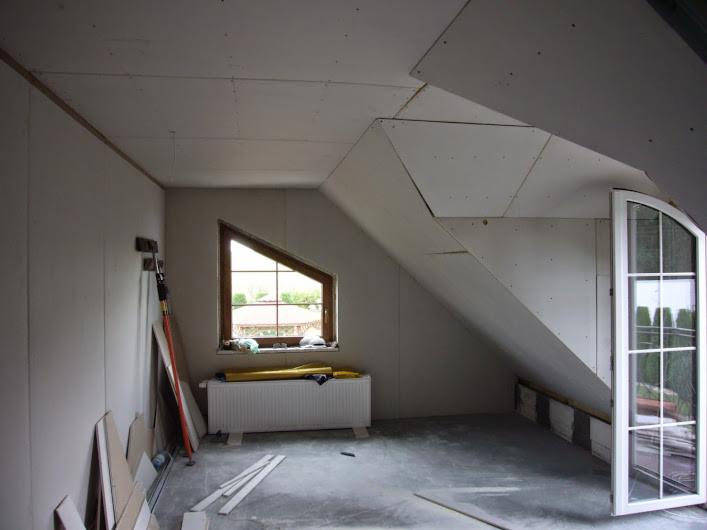 Poddasze w trakcie prac wykończeniowych: płyty gipsowe na ścianach i suficie, okno trójkątne, drzwi balkonowe. Widoczne narzędzia i materiały budowlane.