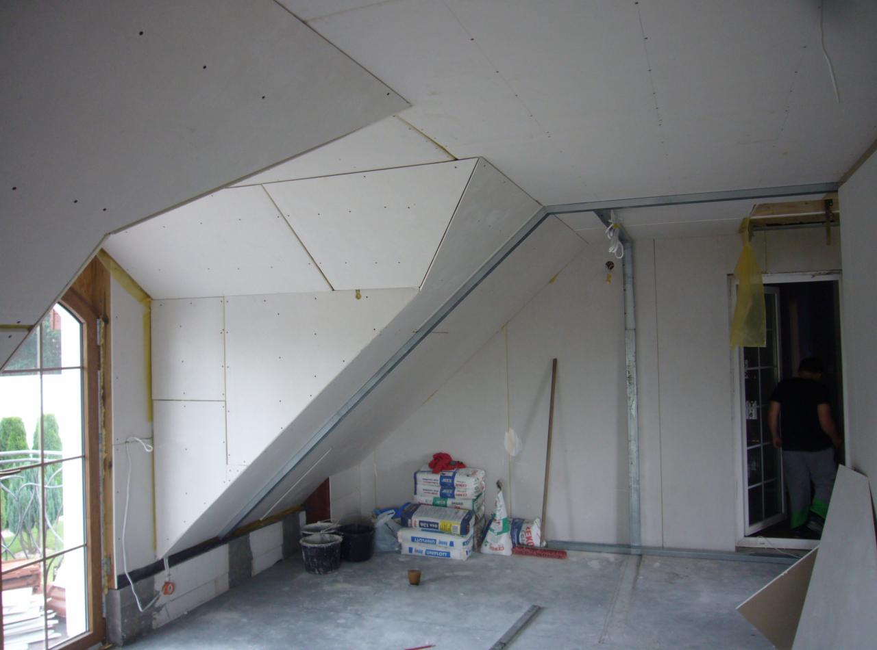 Poddasze w trakcie zabudowy płytami gipsowo-kartonowymi. Widoczne profile, worki z zaprawą i okno. Prace wykończeniowe w surowym stanie.