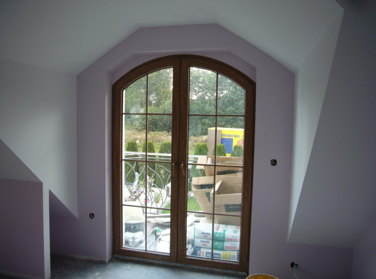 Poddasze z oknem balkonowym w drewnianej ramie po malowaniu ścian na jasny fiolet. Widoczne materiały budowlane i narzędzia.