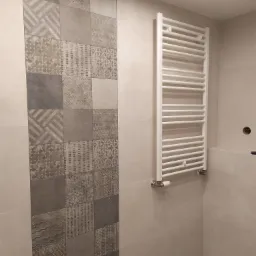 Wykończona łazienka z dekoracyjną ścianą z płytek w odcieniach szarości i beżu oraz białym grzejnikiem drabinkowym.