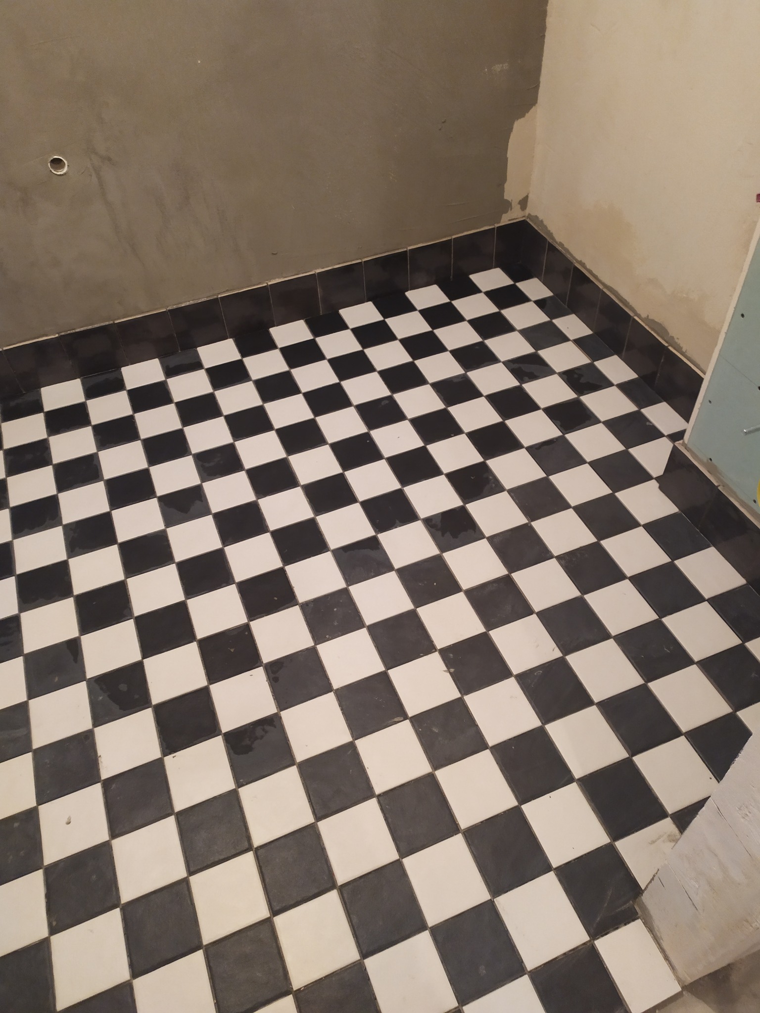 Ułożona podłoga z czarno-białych kwadratowych płytek w trakcie remontu łazienki, ściany częściowo wykończone szarym tynkiem, widoczne niedoskonałości i brak fug.
