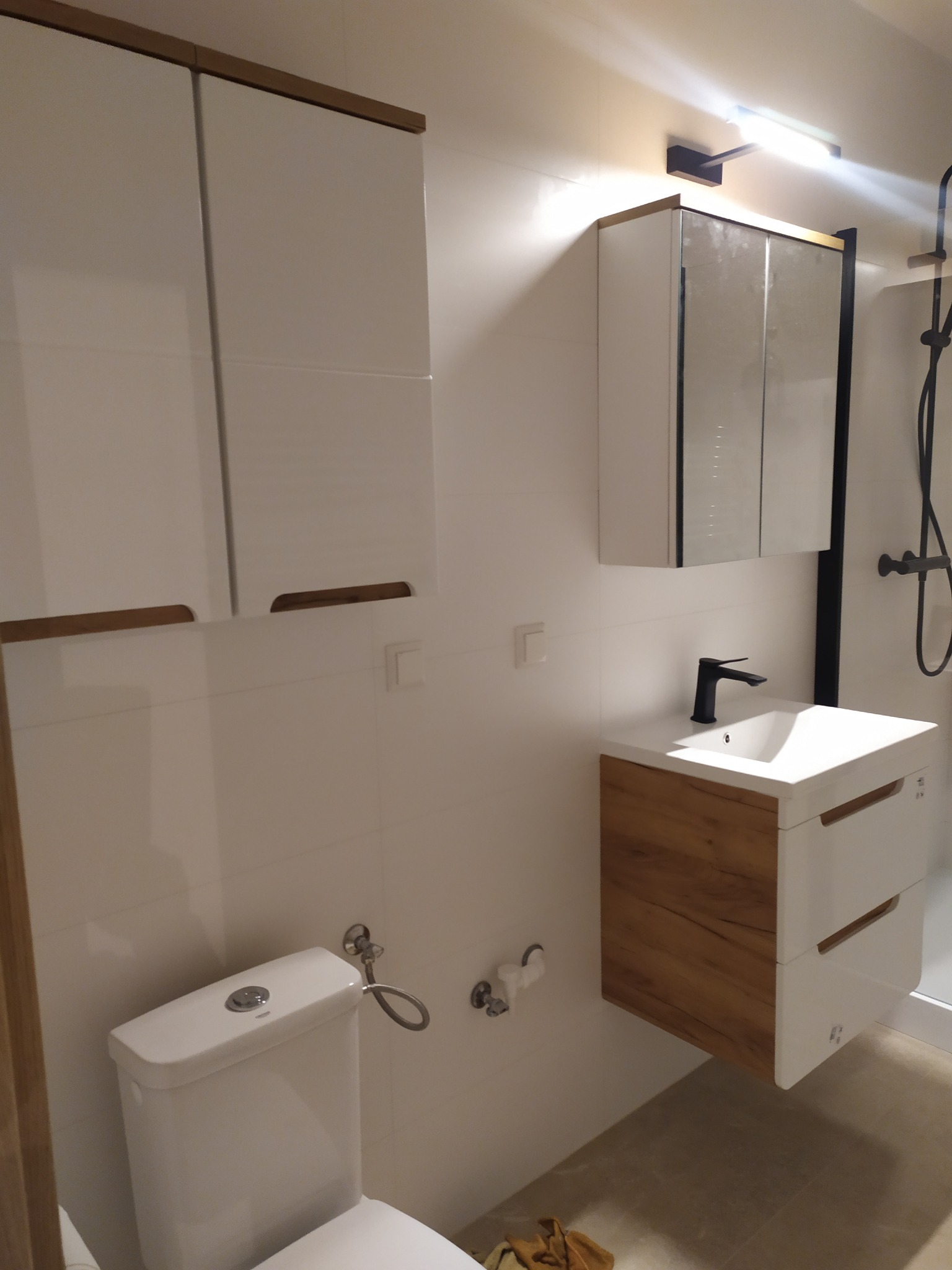 Nowoczesna łazienka z białymi płytkami, wiszącą szafką z lustrem, umywalką z drewnopodobną szafką i czarną armaturą oraz toaletą z bidetką.