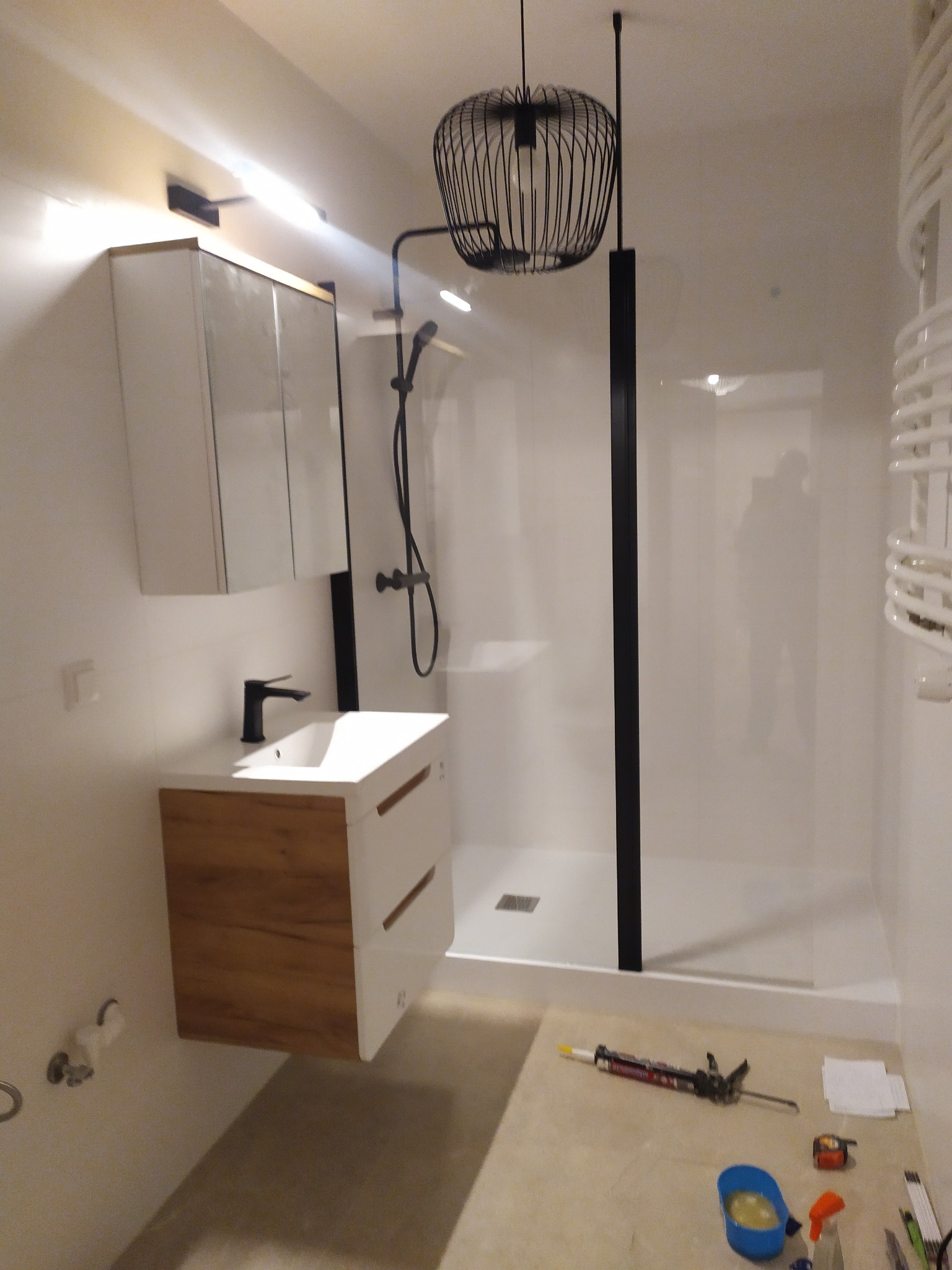 Nowoczesna łazienka z kabiną prysznicową walk-in z czarnymi profilami, umywalką z szafką z drewnianym frontem, czarną armaturą i dekoracyjną lampą wiszącą. Na podłodze leżą narzędzia i akcesoria...