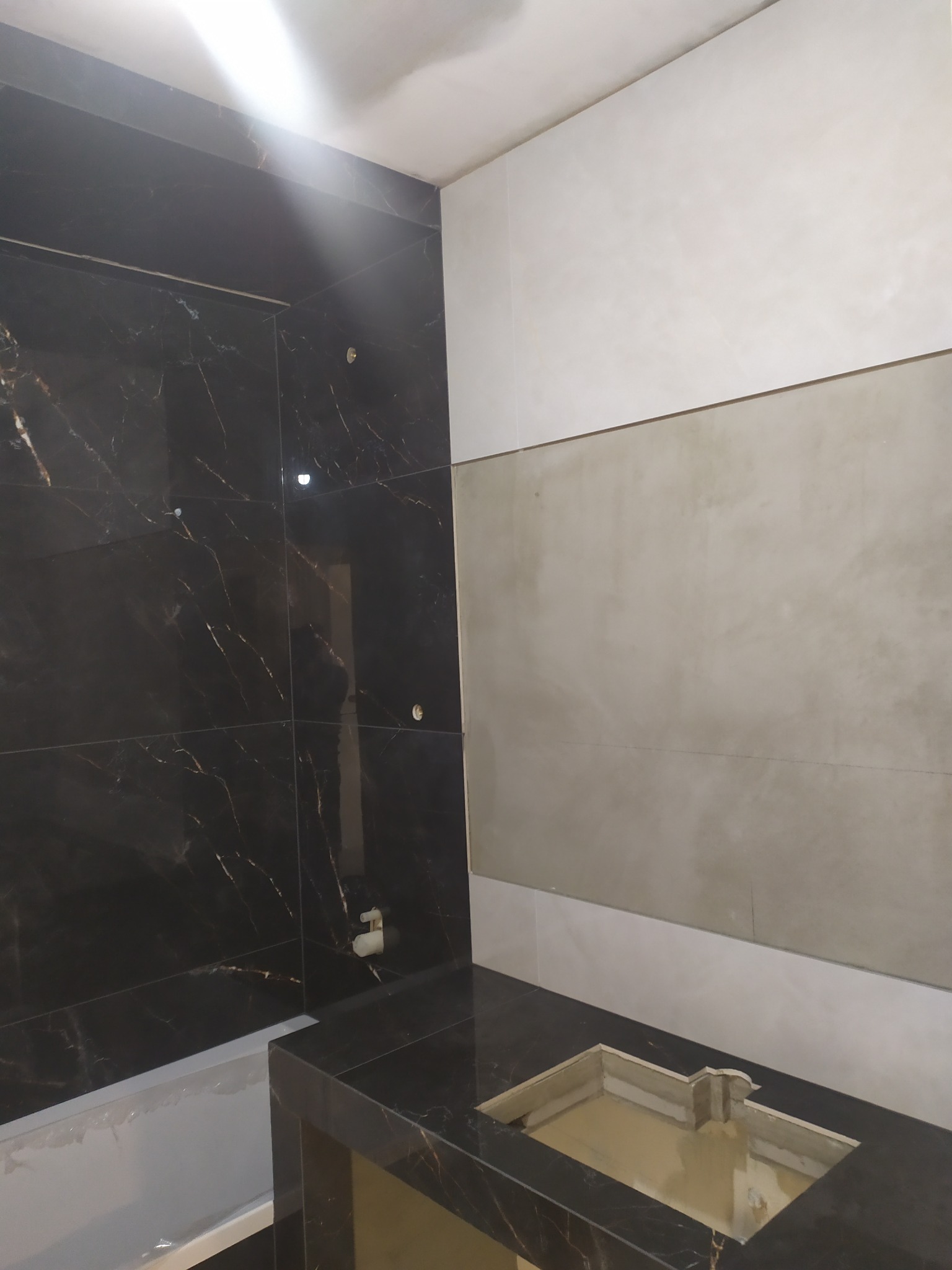 Częściowo wykończona łazienka z czarnymi płytkami imitującymi marmur na ścianie i blacie umywalkowym, oraz jasnymi płytkami na pozostałej części ściany.