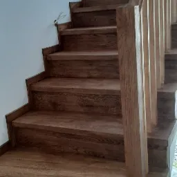 Drewniane schody o ciemnym odcieniu drewna, z widoczną strukturą słojów, prowadzące w górę wzdłuż białej ściany. Widoczne niedociągnięcia przy ścianie sugerują prace wykończeniowe.