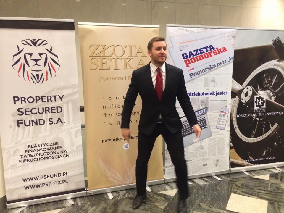 Mężczyzna w garniturze na tle roll-upów z logo firm finansowych i Gazety Pomorskiej. Prezentacja oferty finansowej, być może kredytowej.
