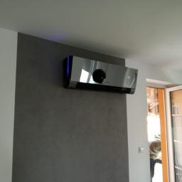 A-Z Serwis Włodzimierz Stasiuk - Nowoczesny klimatyzator w kolorze srebrnym, zamontowany na szarej ścianie w minimalistycznym wnętrzu, widoczny fragment otwartych drzwi balkonowych.