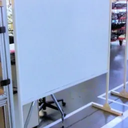 Biały, mobilny parawan spawalniczy na hali produkcyjnej, z metalową ramą i widocznymi elementami konstrukcyjnymi. Podłoga z wyznaczonymi strefami.