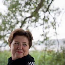 Portret uśmiechniętej kobiety w plenerze, z drzewem w tle. Ubrana w bluzkę w paski i czarny szal. Złote kolczyki, krótka fryzura. Spokojny wyraz twarzy.