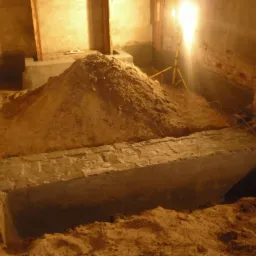 Wykop pod fundament z widocznym zbrojeniem i częściowo wylanym betonem, oświetlony lampą budowlaną na trójnogu, w tle ceglana ściana.