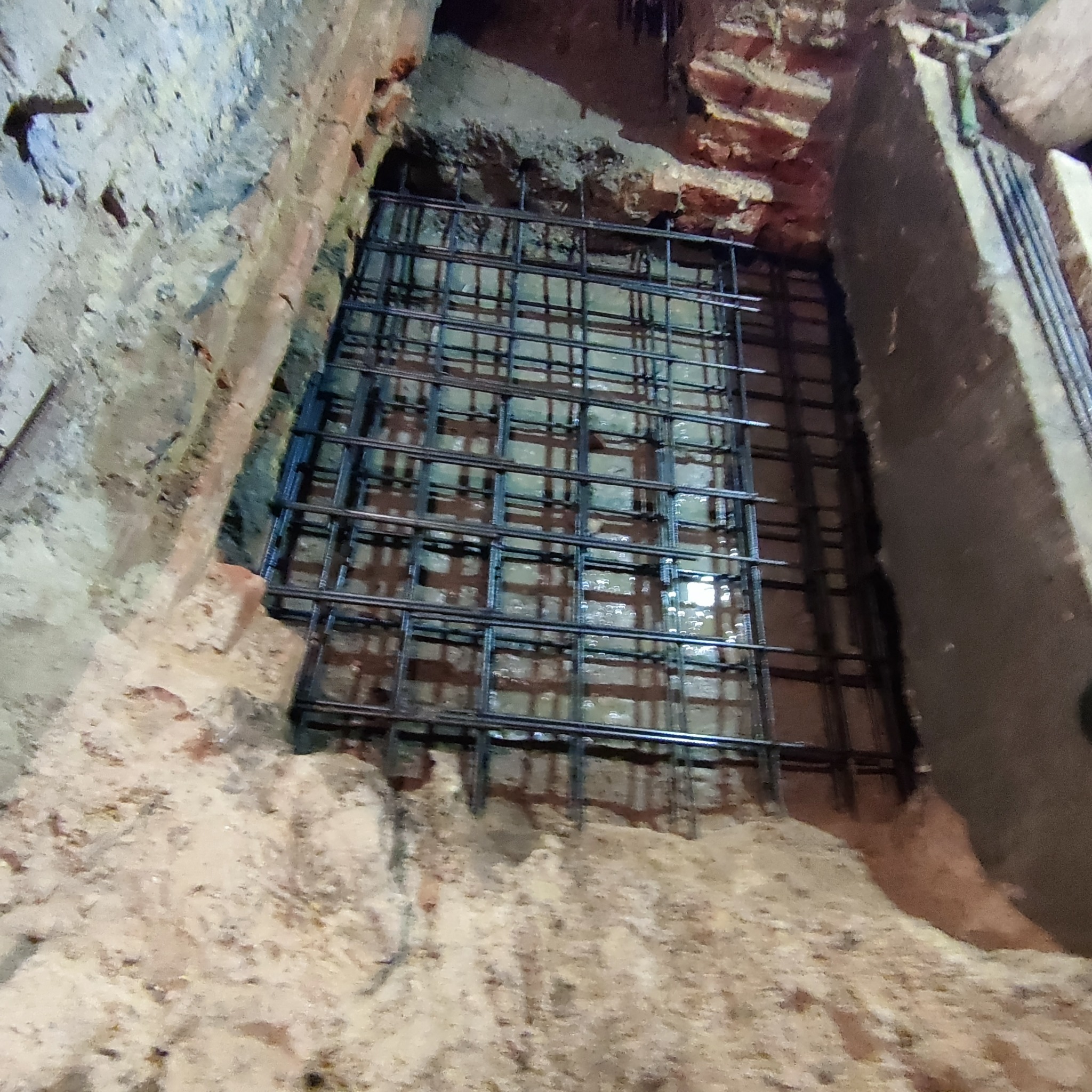 Zbrojenie w wykopie fundamentowym, widoczne pręty stalowe tworzące siatkę, otoczone ziemią i fragmentami cegieł. Przygotowanie do zalewania betonem.