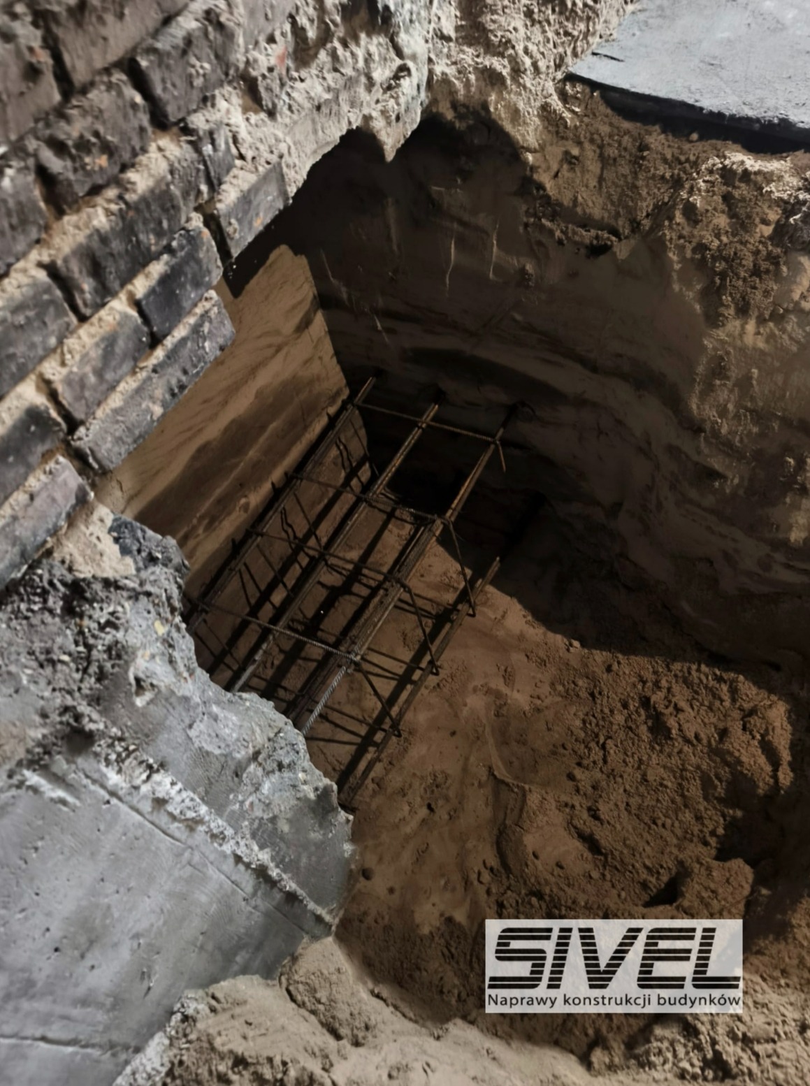 Zbrojenie w wykopie pod fundamenty, widok z góry. Ceglana ściana w tle. Prace ziemne w trakcie realizacji. Logo firmy w rogu kadru.