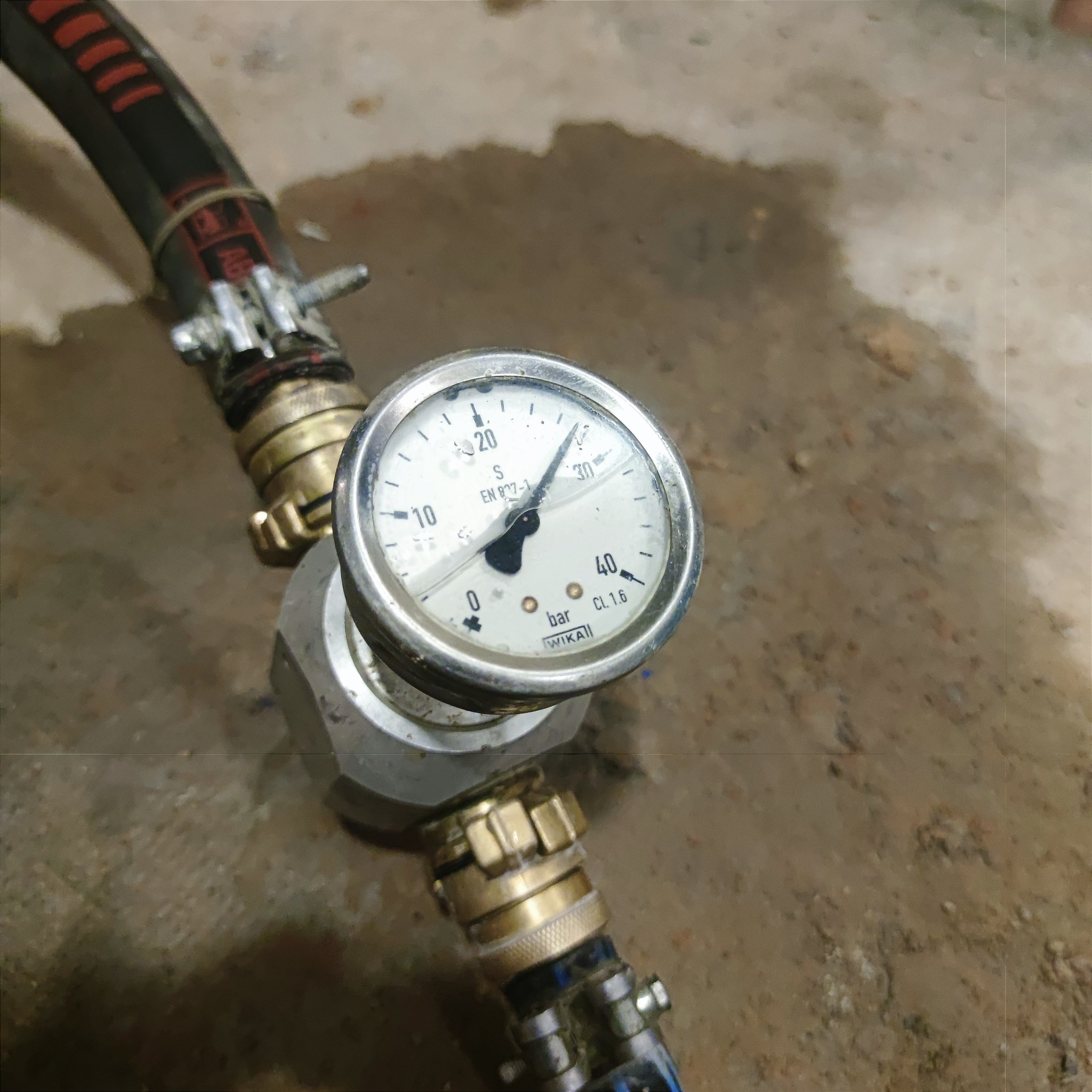 Manometr WIKA z podłączonymi przewodami hydraulicznymi na tle betonowej posadzki. Widoczne złącza i zaciski. Pomiar ciśnienia w instalacji hydraulicznej.