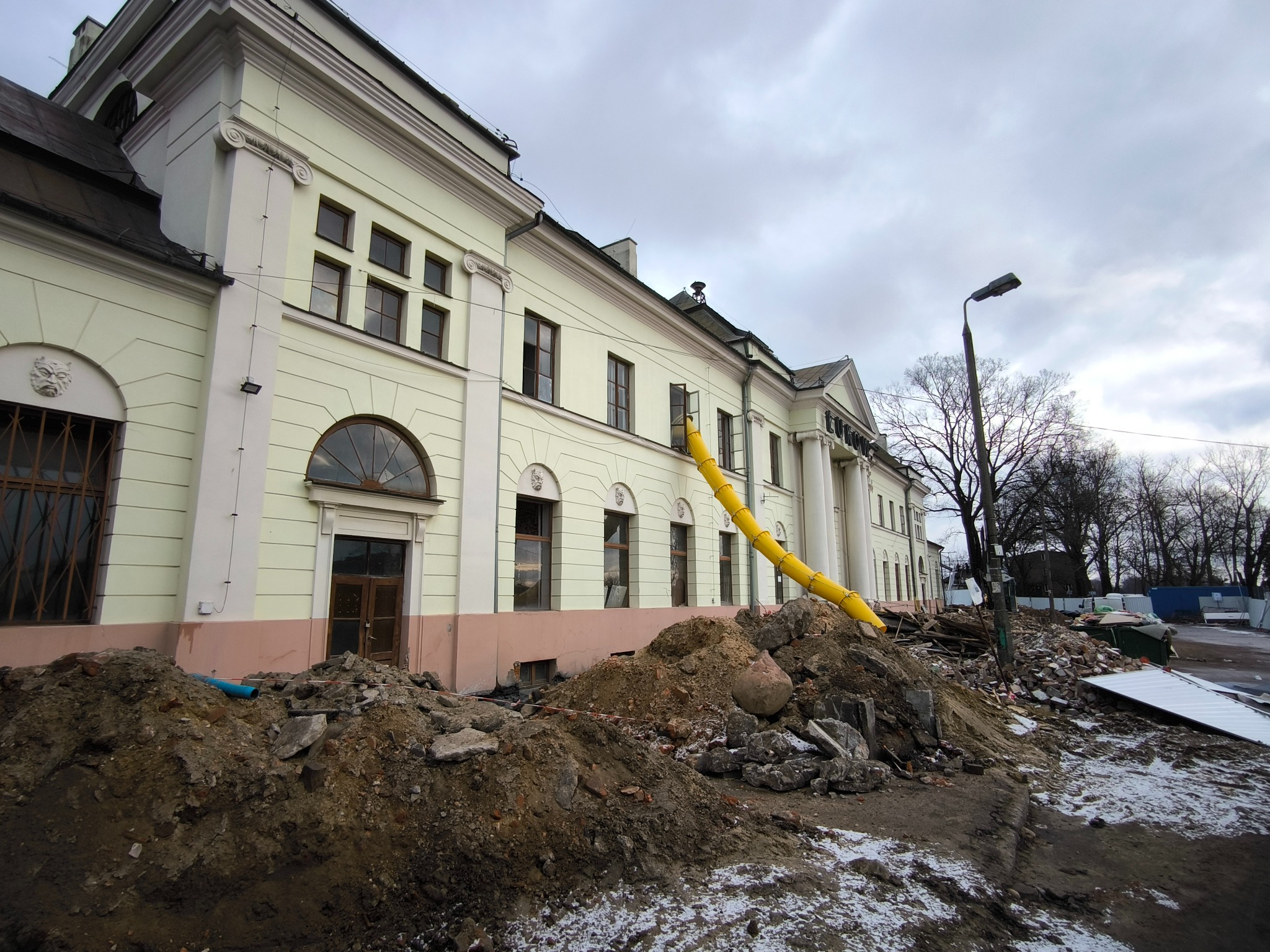 Demontaż budynku z żółtym zsypem gruzu. Stare okna i gruzowisko przed budynkiem. Widok na elewację z kolumnami i detalami architektonicznymi. Pochmurne niebo.