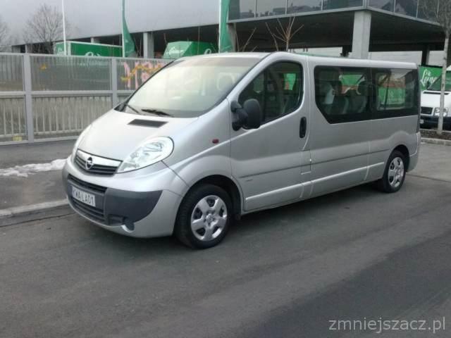 Srebrny Opel Vivaro, bus do przewozu osób, zaparkowany na zewnątrz. Widoczne detale karoserii i felgi. Tło: budynek z przeszkloną fasadą i zielone elementy dekoracyjne.