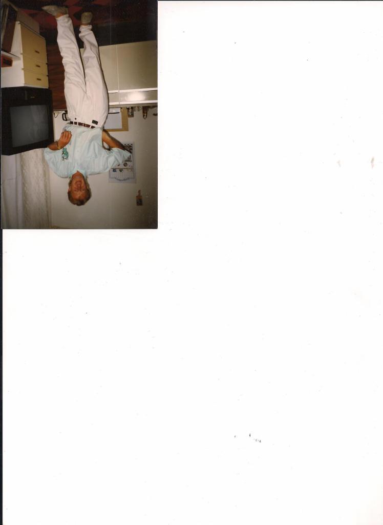 Obrócone o 180 stopni zdjęcie wnętrza kuchni z mężczyzną w jasnych spodniach i koszuli. Widoczny telewizor, szafki i kalendarz na ścianie.