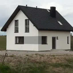 Nowoczesny, biały dom z czarnym dachem z blachodachówki, szarym pasem elewacji, małymi oknami i czarnym kominem, widziany z poziomu gruntu.