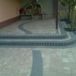 Wykończony taras z kostki brukowej w odcieniach beżu i szarości, z dekoracyjnymi schodami i obrzeżami. Widoczny grill i roślinność w tle.