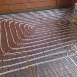 Białe rury ogrzewania podłogowego ułożone na folii z siatką na tle ceglanej ściany. Widoczny fragment opakowania materiałów.