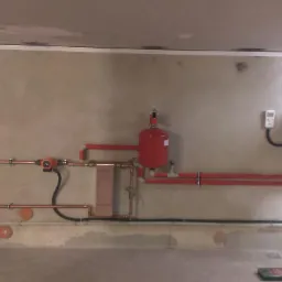 Zainstalowany system grzewczy z czerwonym zbiornikiem wyrównawczym, miedzianymi rurami i czarną skrzynką sterowniczą na surowej ścianie w pomieszczeniu gospodarczym.