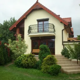 Dwupiętrowy dom z beżową elewacją, balkonem z ozdobną, czarną balustradą i schodami wejściowymi otoczony zielenią.