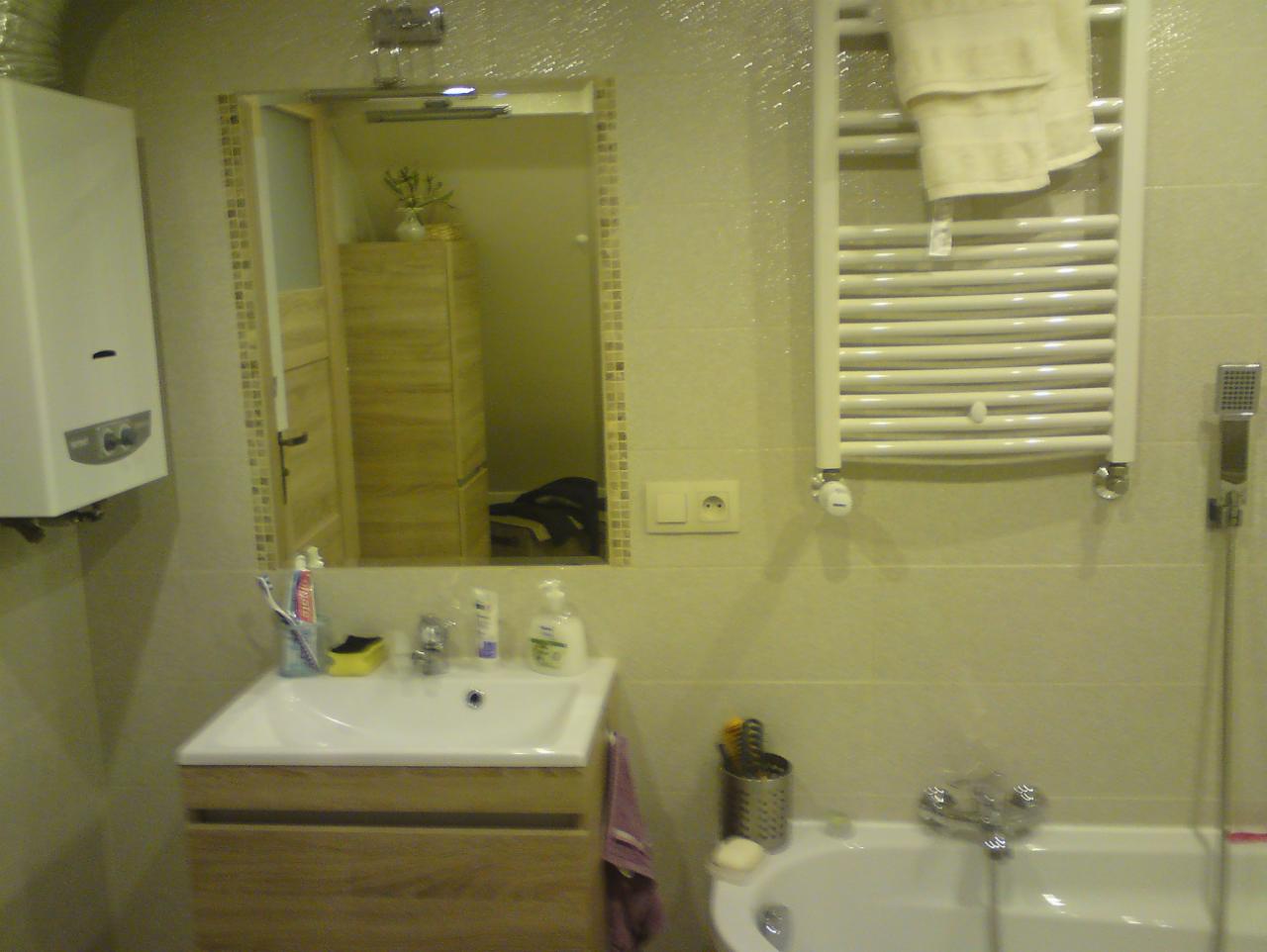 Jasna łazienka po remoncie: umywalka z szafką, grzejnik drabinkowy z ręcznikiem, wanna i lustro z mozaikową ramą. Widoczny bojler i akcesoria łazienkowe.