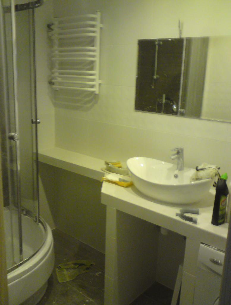 Wykończona łazienka z kabiną prysznicową, umywalką nablatową i grzejnikiem drabinkowym. Widoczne narzędzia i środki czystości. Oświetlenie górne.