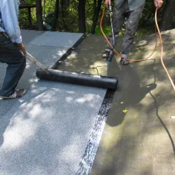 Dekarze podczas uszczelniania dachu papą termozgrzewalną przy użyciu palnika i wałka. Widoczne fragmenty dachu pokryte papą i narzędzia pracy.