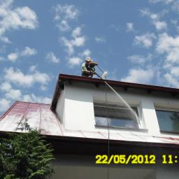 Malowanie Dachów Sławomir Jankowski - Pracownik czyści dach z okiennic za pomocą myjki ciśnieniowej. Widoczne białe chmury na niebie i fragment zielonego drzewa. Ujęcie z dołu.