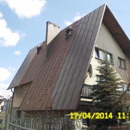 Malowanie Dachów Sławomir Jankowski - Dwaj pracownicy na drabinach malują blaszany dach domu jednorodzinnego o nietypowej architekturze, z widocznym kominem i datą na pierwszym planie.