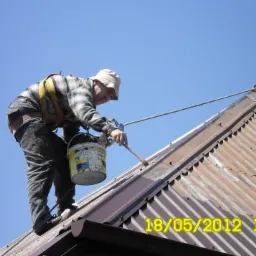 Pracownik z zabezpieczeniem maluje rdzawy, blaszany dach brązową farbą. Widoczny fragment dachu, niebieskie niebo i data nałożona na zdjęcie: 18/05/2012.