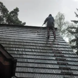 Pracownik na drabinie czyści dach z drewnianych gontów za pomocą myjki ciśnieniowej. Widok z dołu, pochmurne niebo w tle.