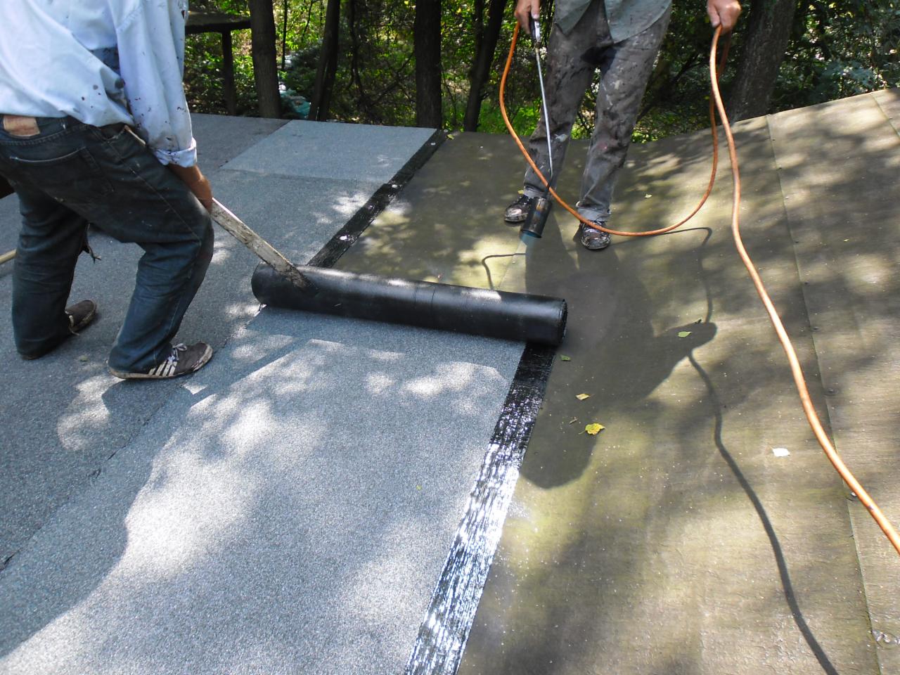 Dekarze podczas uszczelniania dachu papą termozgrzewalną przy użyciu palnika i wałka. Widoczne fragmenty dachu pokryte papą i narzędzia pracy.