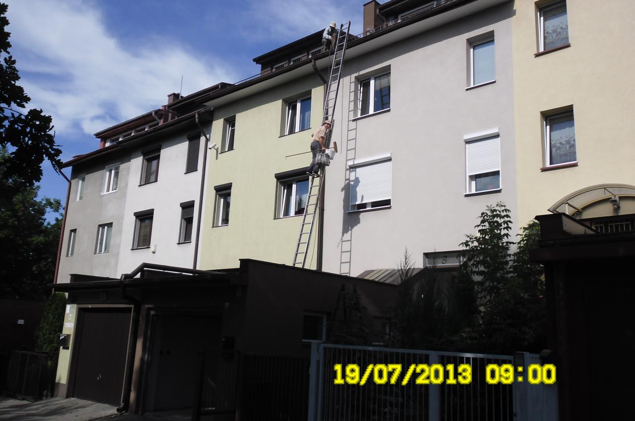 Dwóch mężczyzn na drabinie przy budynku, jeden na dachu, drugi maluje elewację. Widoczne rynny i okna z roletami. Ujęcie z dołu, data 19/07/2013.