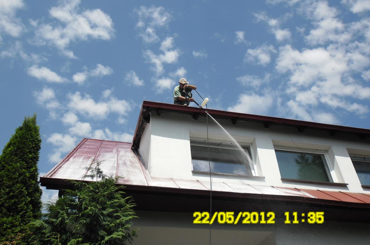 Pracownik czyści dach z okiennic za pomocą myjki ciśnieniowej. Widoczne białe chmury na niebie i fragment zielonego drzewa. Ujęcie z dołu.