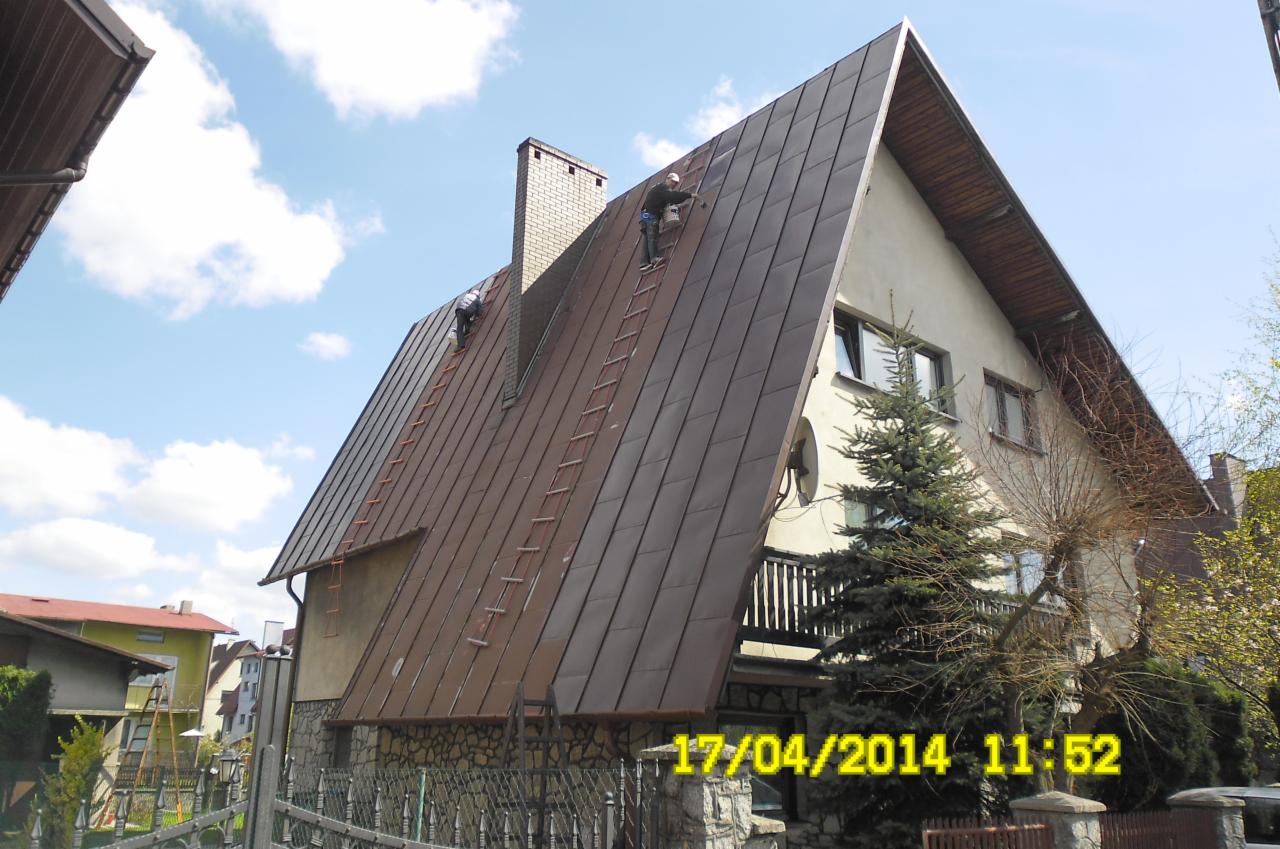 Dwaj pracownicy na drabinach malują blaszany dach domu jednorodzinnego o nietypowej architekturze, z widocznym kominem i datą na pierwszym planie.