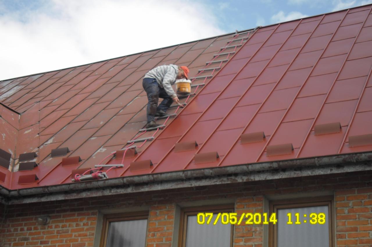 Pracownik na drabince maluje czerwony dach blaszanego budynku. Widoczny fragment muru i okna. Praca na wysokości z użyciem drabiny dekarskiej.