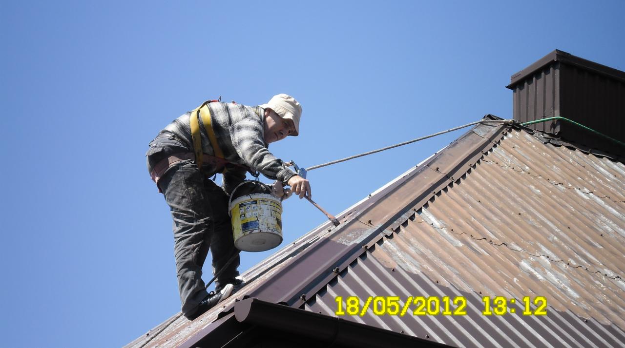 Pracownik z zabezpieczeniem maluje rdzawy, blaszany dach brązową farbą. Widoczny fragment dachu, niebieskie niebo i data nałożona na zdjęcie: 18/05/2012.