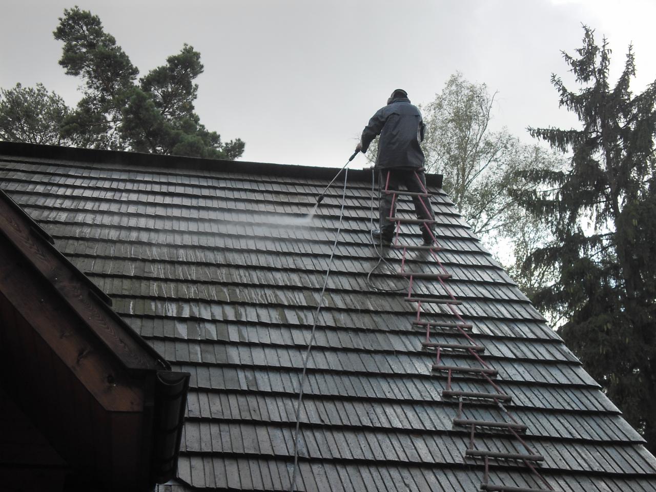 Pracownik na drabinie czyści dach z drewnianych gontów za pomocą myjki ciśnieniowej. Widok z dołu, pochmurne niebo w tle.