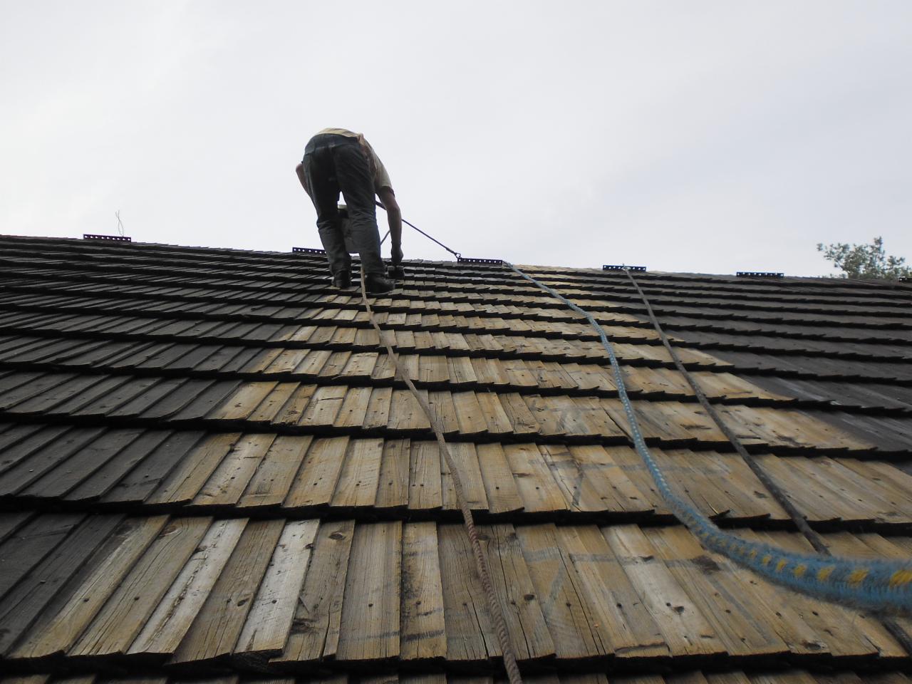 Pracownik zabezpieczony linami na stromym dachu krytym drewnianym gontem. Widok z dołu na osobę wykonującą prace na wysokości.
