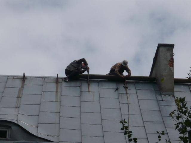 Dwóch dekarzy na blaszanym dachu w Łasku, jeden wierci wiertarką, drugi używa młotka. Widoczny komin i pochmurne niebo w tle.