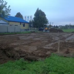 Przygotowanie terenu pod budowę: wyrównana ziemia, widoczne zbrojenie i szalunki. W tle budynek z niebieskim dachem i sprzęt budowlany.