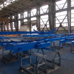 Niebieskie, pomalowane proszkowo metalowe konstrukcje na stojakach w hali produkcyjnej z dużymi oknami i stalową konstrukcją dachu.