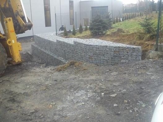 Gabiony w ogrodzie, ułożone schodkowo, z żółtą koparką w tle. Widoczne trawnik i budynek. Konstrukcja z kamieni w metalowych koszach.