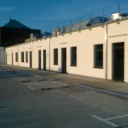 Długi budynek z białymi ścianami i ciemnymi oknami, widoczny z poziomu parkingu z elementami spowalniającymi ruch.