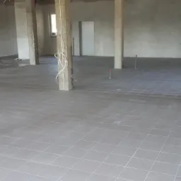 Duża powierzchnia z ułożonymi szarymi płytkami podłogowymi w surowym stanie, widoczne betonowe kolumny i wystające rury instalacyjne.