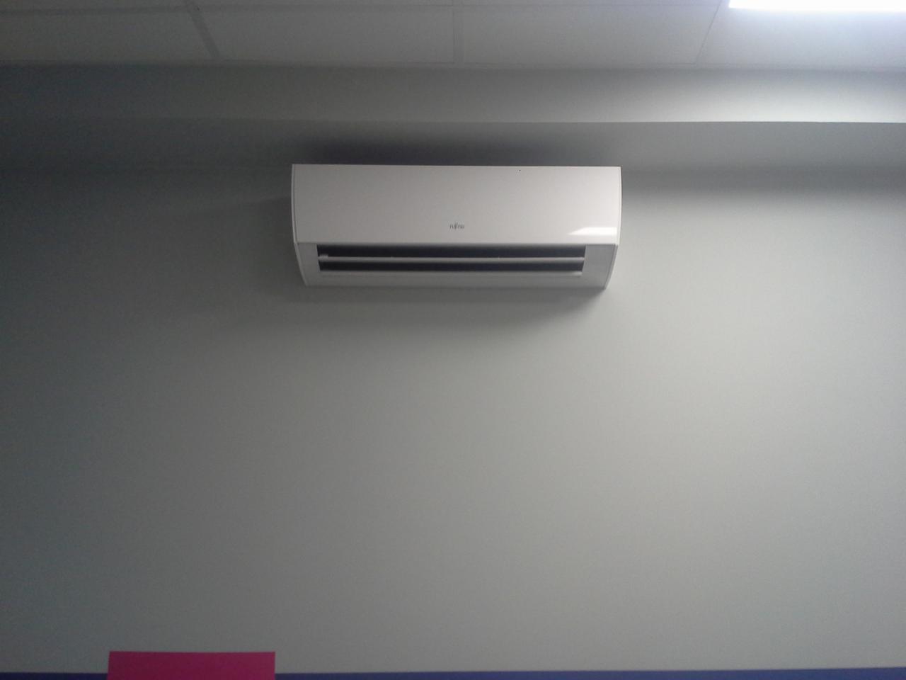 Biały klimatyzator Fujitsu zamontowany na szarej ścianie w biurze, widoczny fragment sufitu i listwy przypodłogowej.