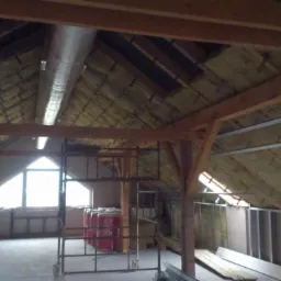 Poddasze w trakcie adaptacji: widoczne drewniane belki, ocieplenie ścian i dachu wełną, rusztowania oraz okno w szczycie. Prace wykończeniowe.