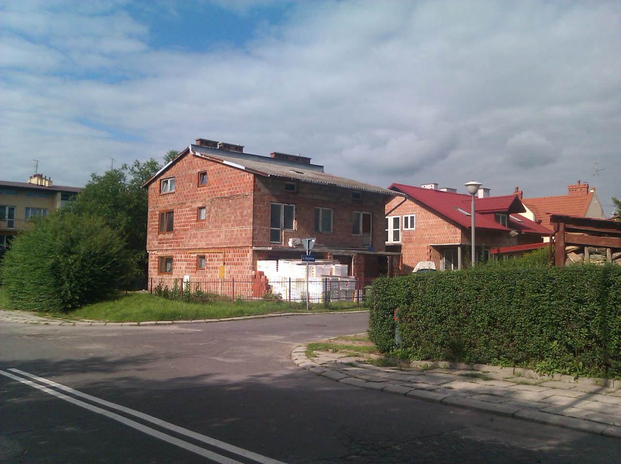 Niedokończony, dwupiętrowy budynek z czerwonej cegły z widocznymi oknami, z materiałami budowlanymi składowanymi na zewnątrz, w tle inne domy i pochmurne niebo.